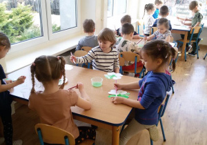 Widok na siedzące przy stolikach dzieci, które malują zieloną farbą czterolistną koniczynkę.